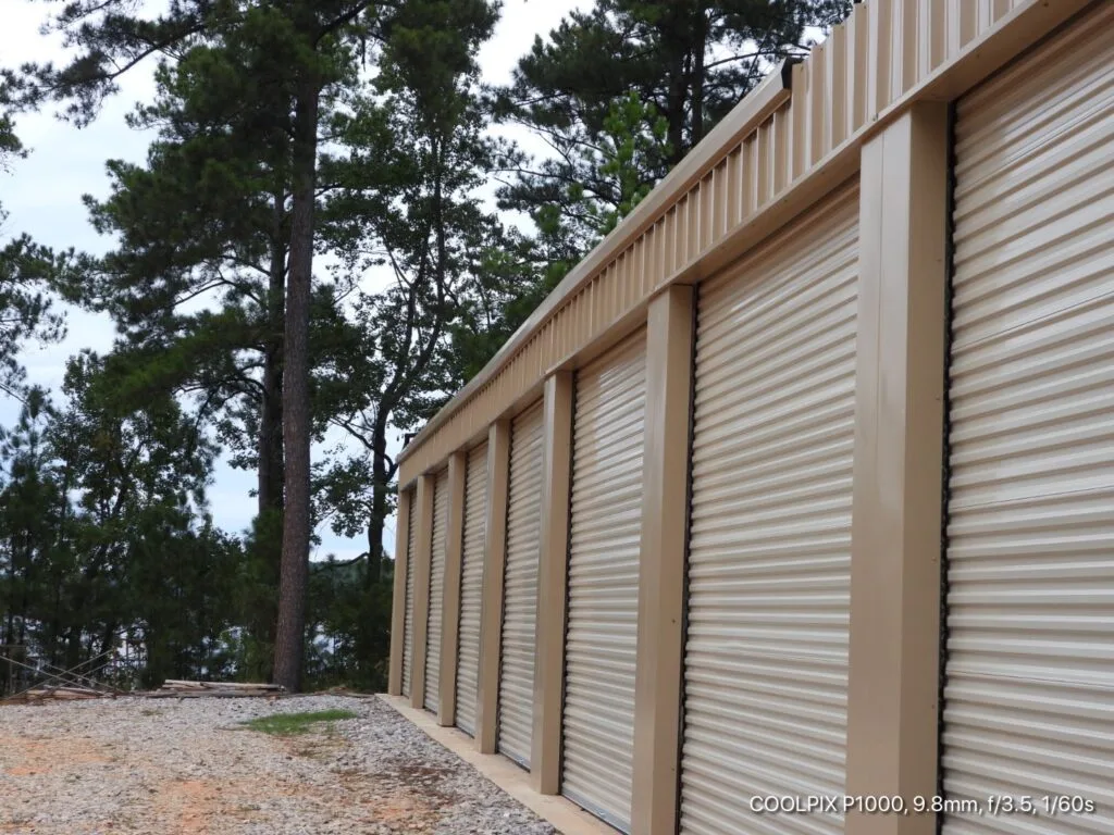 Beige self-storage units with roll-up doors line a dirt path bordered by tall pine trees.